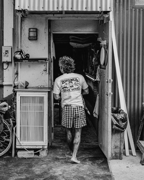 A person with tousled hair, wearing the Golden Hawaii T-Shirt in Vintage White and checkered shorts, walks barefoot into a cluttered room through an open door. The black and white scene looks like it's set in an alley or workshop.