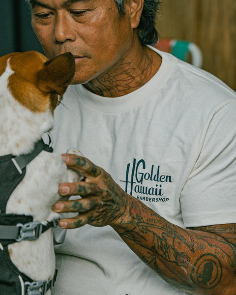 A tattooed man wears a white Golden Hawaii T-Shirt - Arctic Blue and gently holds a small, harnessed dog facing him in a calm, affectionate scene.