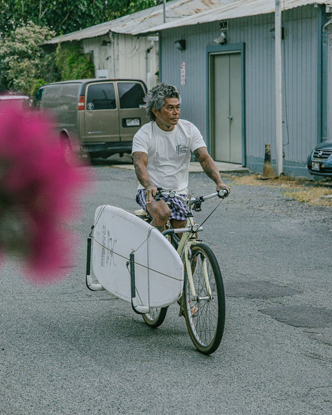 A person with gray hair rides a bike down the street, carrying a white surfboard in a side rack. They wear the Golden Hawaii T-Shirt - Arctic Blue and plaid shorts. A van and gray building are behind them, with pink flowers in front.
