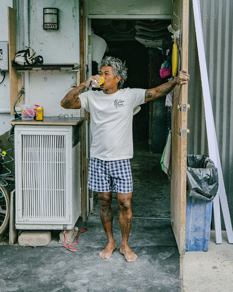 A barefoot, gray-haired person with tattoos stands in a doorway wearing the Golden Hawaii T-Shirt in Arctic Blue and plaid shorts, sipping from a can. One hand holds the door, creating a laid-back atmosphere.