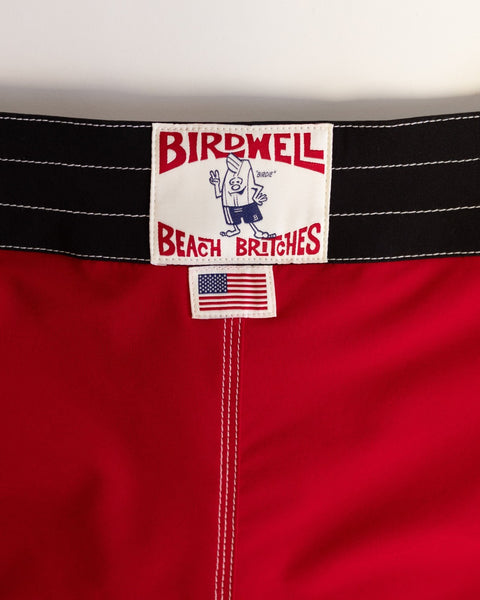 Close-up of 808 Mono Stripe - Red/Black boardshort waistband with black and white stitching, featuring a Birdwell Beach Britches label with cartoon bird and American flag tag—proudly made in USA.
