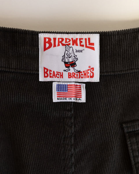 A close-up of the corduroy waistband on the Classic Corduroy Shorts - Oregano, showing two labels: one with Birdwell Beach Britches and a cartoon bird, and another featuring an American flag with 