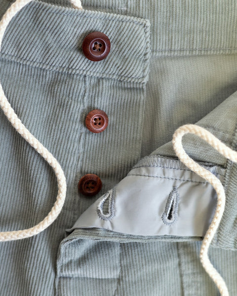Close-up of Classic Corduroy Shorts - Eucalyptus: light green corduroy, three brown buttons, white adjustable drawcord waist, and visibly stitched buttonholes on light gray fabric lining.