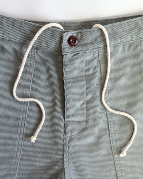 Close-up of Classic Corduroy Shorts - Eucalyptus in light green, featuring a brown button and adjustable drawcord waist, loosely tied at the waistband, shown against a plain white background.