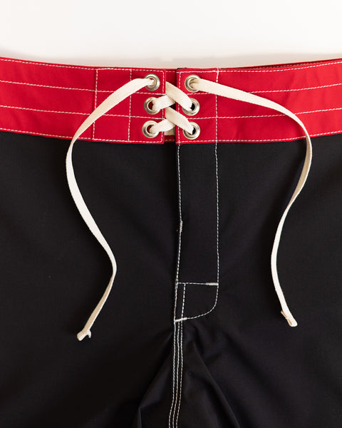 Close-up of the waistband of the 808 Mono Stripe - Black/Red boardshort, made in USA, showing a red top band, white drawstrings through metal eyelets, and white stitching along the seams.