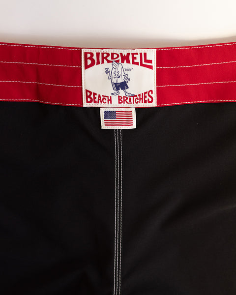 Close-up of medium-length 808 Mono Stripe - Black/Red SurfStretch boardshorts with a red waistband, white stitching, Birdwell Beach Britches label with cartoon bird, and a small American flag patch. Made in USA.