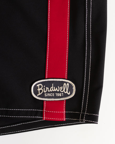 Close-up of 808 Mono Stripe - Black/Red SurfStretch boardshort fabric, showing a vertical red stripe, white stitching, and a Birdwell Since 1961 oval patch near the hem. Made in USA.