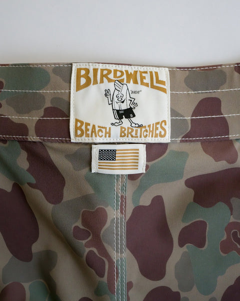 Close-up of Frogskin Camo 808 SurfStretch Side Pocket Boardshorts featuring a white Birdwell Beach Britches label with a cartoon character and a small American flag patch, highlighting these water-repellent boardshorts made in the USA.