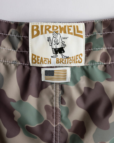 Close-up of 300 Boardshorts - Frogskin Camo, made in USA, featuring a Birdwell Beach Britches label with a cartoon character and a small embroidered American flag patch below the waistband.