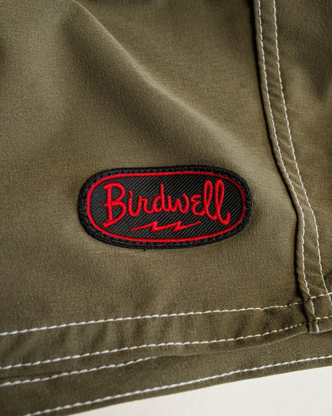 Close-up of the dark olive 808 Powerbird SurfStretch boardshort fabric with white stitching and a black and red Birdwell patch featuring a lightning bolt design below the cursive text.