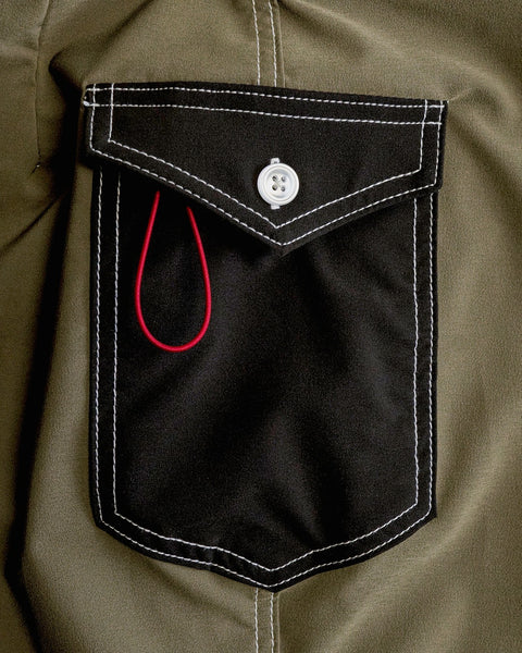 A close-up of the 808 Powerbird - Dark Olive boardshort pocket shows white stitching, a white button, and a red thread loop on a black pocket set against a dark olive fabric background.