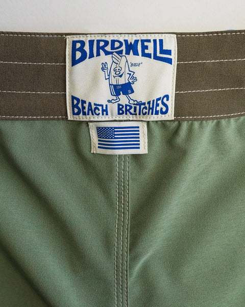 Close-up of a tag on olive 808 Powerbird SurfStretch swim trunks. The tag reads Birdwell Beach Britches in blue with a cartoon mascot and features an American flag patch, showing these stretch boardshorts are made in the USA.
