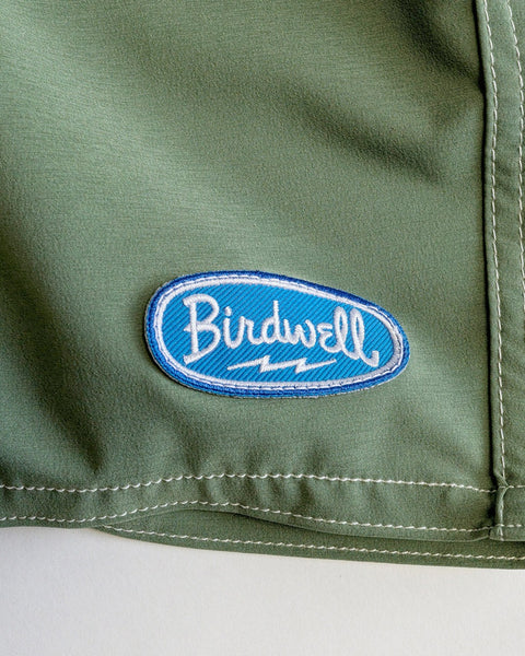 Close-up of the Olive 808 Powerbird's SurfStretch fabric with a blue and white oval Birdwell patch stitched near the seam, highlighting iconic USA-made quality.