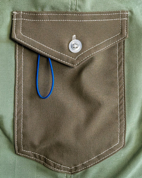 A close-up of the olive 808 Powerbird shirt pocket with white stitching. A blue SurfStretch band, inspired by boardshort tech and made in the USA, is partially tucked in, hanging over the edge.