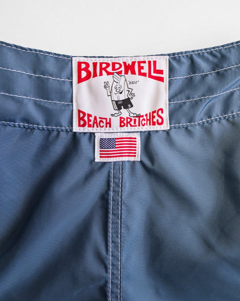 A close-up of the 400 Boardshorts - Federal Blue waistband shows the Birdwell Beach Britches label with a cartoon character and a small American flag tag, highlighting its made in USA quality.
