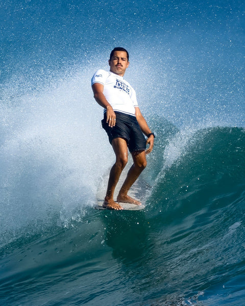 A man wearing a white shirt and navy 300 Boardshorts surfs confidently on a wave, ocean spray around him beneath a clear blue sky.
