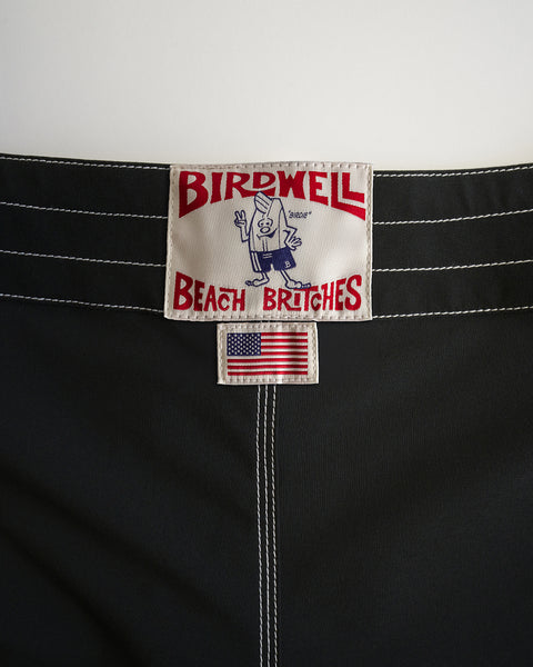 Close-up of a label on 808 Racing Stripes - Black/Olive boardshorts. The top label shows Birdwell Beach Britches with a cartoon bird in swimwear, and a small American flag patch below it. White stitching accents the fabric.