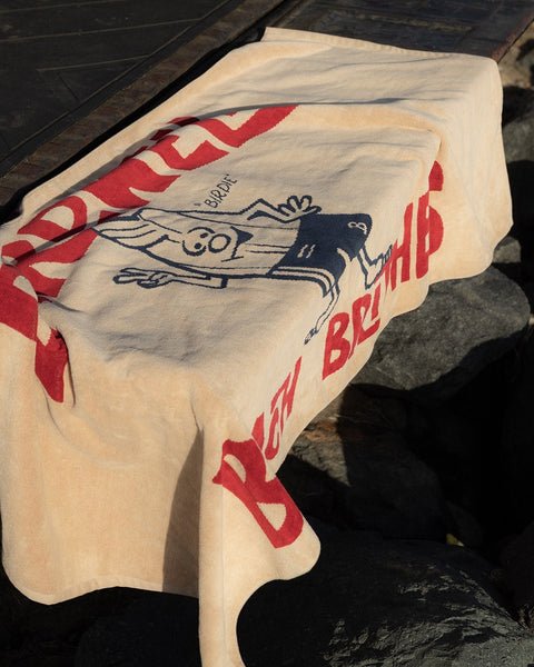 The License Plate Towel - Bone is a 100% cotton, over-sized beige towel featuring red text and a cartoon-style smiling figure, draped over rocks outdoors. It displays the words BEN BRUNS, blending comfort with playful design.
