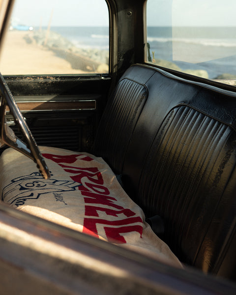 A vintage car with black leather seats and a steering wheel has the License Plate Towel - Bone, featuring red text and a cartoon face, draped over the front seat. The beach and ocean can be seen outside the window.