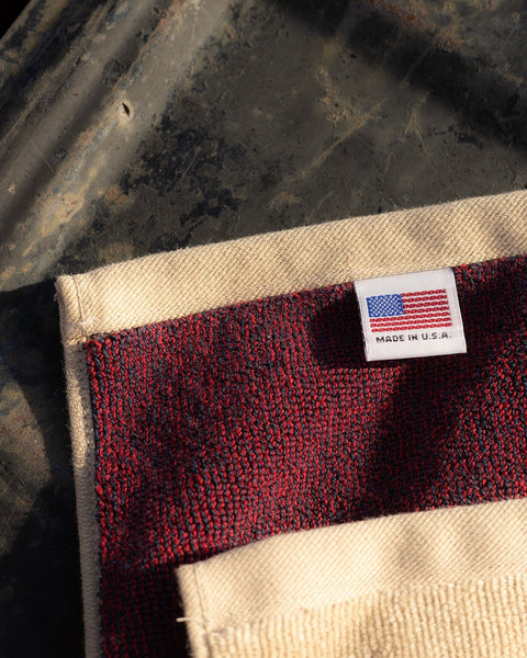A close-up of the License Plate Towel - Bone, an oversized 100% cotton towel with red and cream stripes, a sewn American flag label marked MADE IN U.S.A. in the corner, shown on a weathered metal surface.