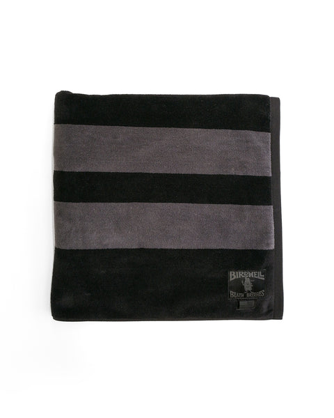 The Comp Stripe Towel - Black/Gray is an oversized, folded black towel featuring two wide dark gray stripes and a Birdwell logo patch at the bottom right corner, made in the USA from 100% cotton and shown on a white background.