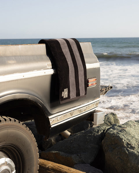 The Comp Stripe Towel - Black/Gray, featuring bold stripes, is draped over a weathered pickup’s tailgate near the ocean, with waves and rocks in the background.