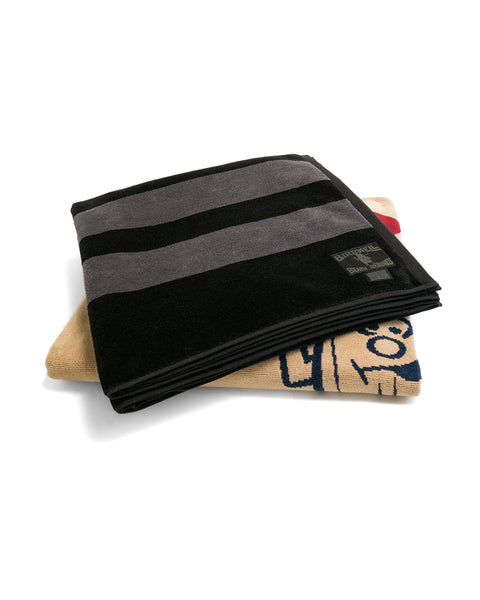 Two folded towels are stacked: the top is the Comp Stripe Towel - Black/Gray (an oversized black towel with gray stripes), and the bottom is tan, featuring part of a cartoon dog’s face and visible red ears.