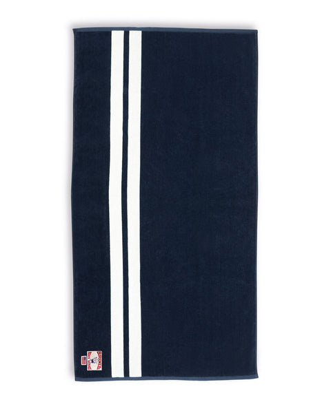 The Comp Stripe Towel - Navy/White is an oversized 100% cotton, navy rectangular towel with two vertical white stripes on one side and a small logo patch in the lower left corner.