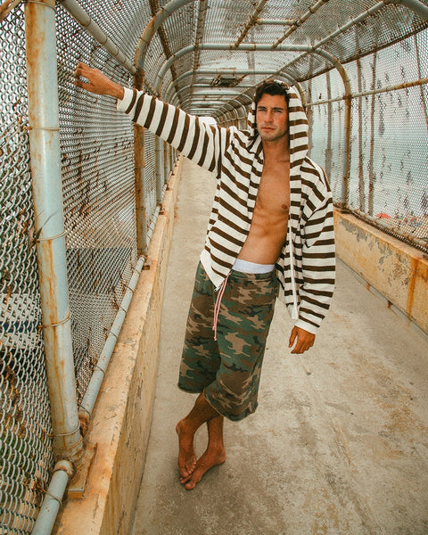 A man in a Zip Hoodie - Vintage White/Brown and camouflage shorts stands barefoot on a fenced pedestrian bridge, leaning against the chain-link fence with one arm extended.