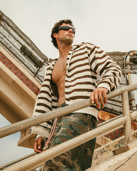 A man wearing sunglasses and a Zip Hoodie - Vintage White/Brown stands outdoors on a staircase with metal railings, looking into the distance. He also wears camouflage pants. The background shows part of a structure.