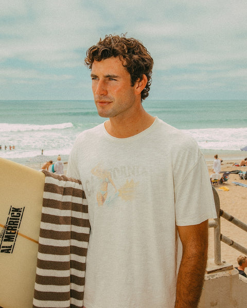 A man with curly brown hair stands by the beach holding a surfboard and a striped towel. He wears the Surf Slub Tee in Vintage White, with the ocean and beachgoers visible in the background.