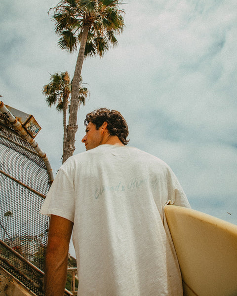 Wearing the Surf Slub Tee in Vintage White, a person stands outdoors by a wire fence and tall palm trees with a surfboard, gazing up at the sky, adding to the sunny, relaxed vibe.