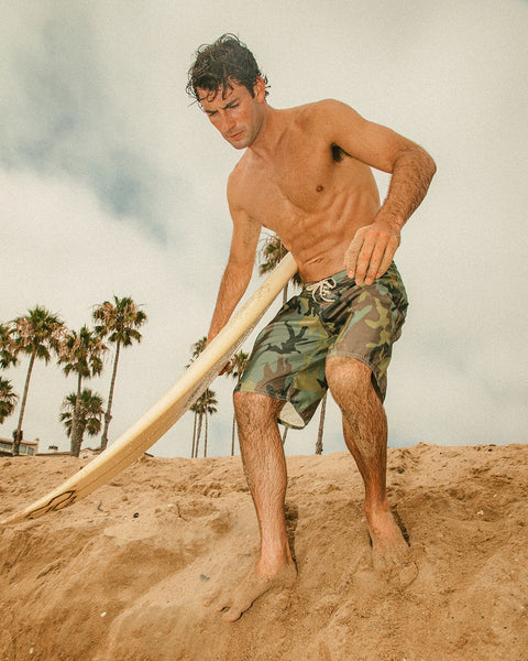 A man in 312 Boardshorts - Woodland Camo, shirtless and barefoot, stands on a sandy beach holding a surfboard as palm trees sway beneath a cloudy sky in the background.