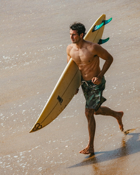 A shirtless man in 312 Boardshorts - Woodland Camo walks barefoot on wet sand, carrying a surfboard under his arm as he heads toward the water.
