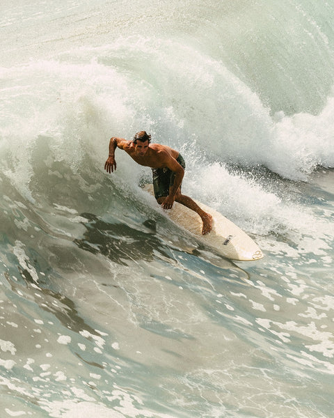 A man with dark hair surfs a large wave, crouched low on his surfboard and wearing Woodland Camo 312 Boardshorts. Ocean water splashes around him as he rides along the curve of the wave.