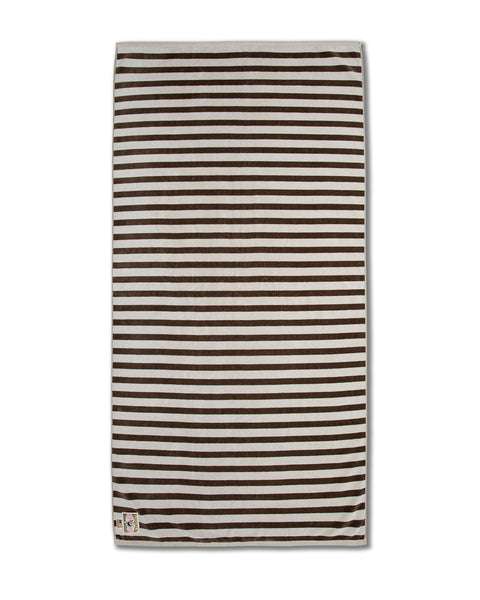 The Beach Towel - Vintage White/Brown is an oversized cotton towel with horizontal brown and white stripes and a small graphic tag on one corner, laid flat on a white background. This limited edition towel offers both style and practicality.