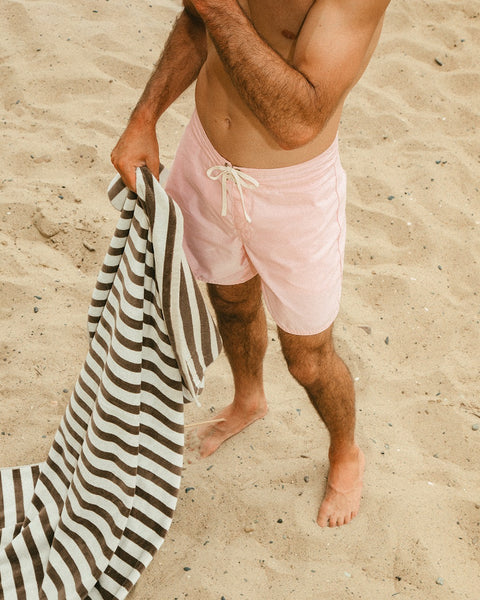 A shirtless person stands on a sandy beach in light pink swim trunks, holding the Beach Towel - Vintage White/Brown. Their head is out of the frame.