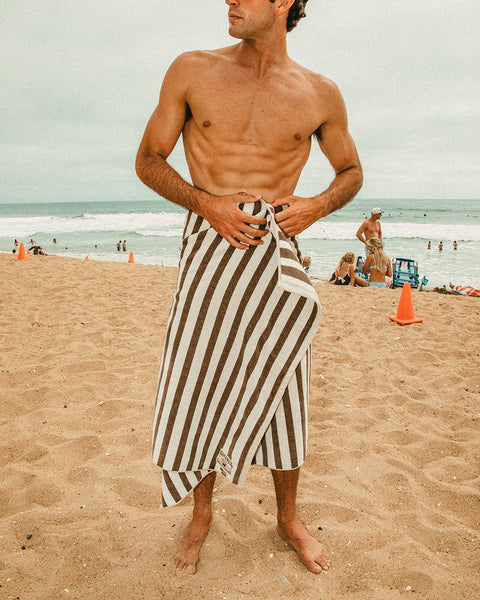 A shirtless man stands on a sandy beach with the limited edition Beach Towel - Vintage White/Brown wrapped around his waist. People and orange cones can be seen in the background by the ocean.