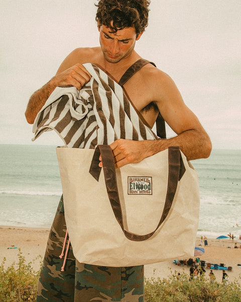 A man at the beach holds a large natural Beach Tote and pulls out a striped towel, with the ocean and sandy shore in the background.