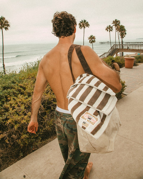 A shirtless man with curly hair walks along a beachside path, carrying a bag with a striped towel and wearing Baggy Raw Edge Short - Woodland Camo. Palm trees, greenery, and the ocean create a relaxed backdrop.