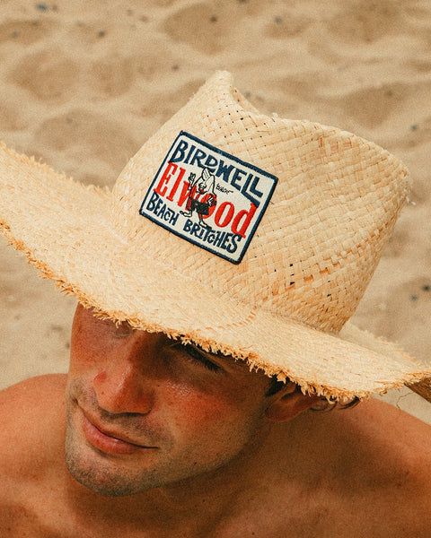 A person wearing the Straw Hat - Natural with a raw edge brim and Birdwell x Elwood Beach Britches patch sits on the sandy beach, their face partly shaded by the wide-brimmed hat.