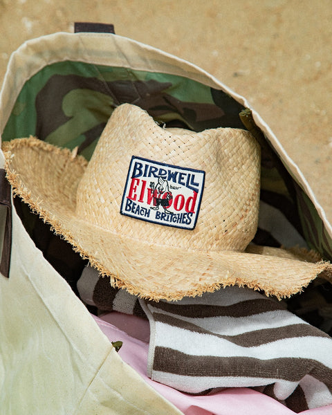 The Straw Hat - Natural with a raw edge brim and signature patch rests on striped towels inside a canvas bag on the sand.