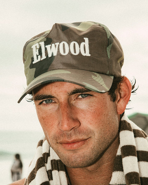A man wearing a Trademark Cap - Woodland Camo and a striped towel around his neck looks into the camera with a neutral expression, set against a beach background.