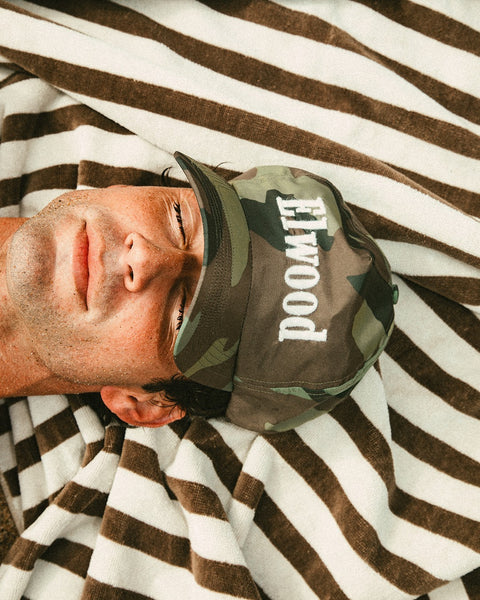 A man lies relaxed on a brown and white striped towel, wearing the limited edition Trademark Cap - Woodland Camo, featuring 
