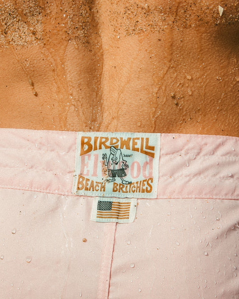 Close-up of a person’s wet back wearing limited edition 310 Boardshorts - Almond Blossom by Birdwell Beach Britches, featuring the iconic cartoon bird label, small American flag, and sand on their skin.