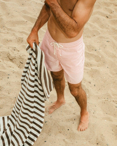 A man stands barefoot on sand, wearing 310 Boardshorts - Almond Blossom and holding a brown and white striped towel. Only his torso and legs are in frame; his face is not visible.