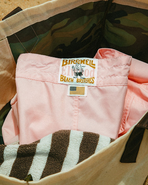 A pair of limited edition 310 Boardshorts - Almond Blossom with a patch and small American flag label rests on a brown and white striped towel inside a canvas bag lined with camouflage.