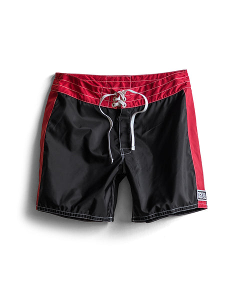 310 Dekka - Black/Red boardshorts with a white drawstring, white stitching, and a small rectangular patch on the bottom left leg, made in the USA and shown on a white background.