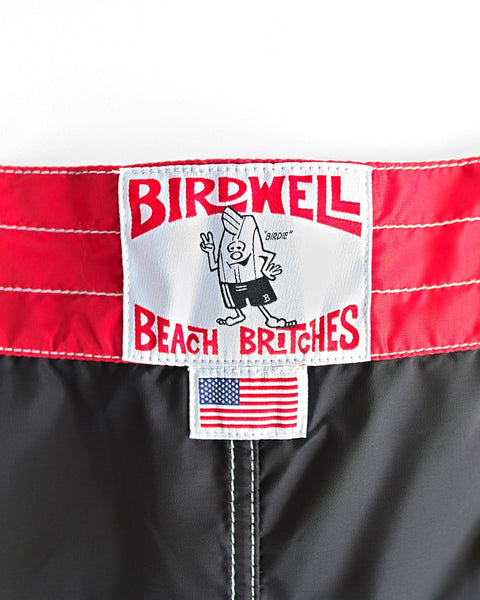 Close-up of a Birdwell 310 Dekka - Black/Red label featuring a cartoon bird in swim trunks, red stitching, and a small American flag tag on black boardshorts made in the USA.