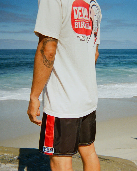 Wearing a white graphic tee and 310 Dekka - Black/Red boardshorts, a person stands on a sandy beach by the ocean, gentle waves rolling in under a clear blue sky.
